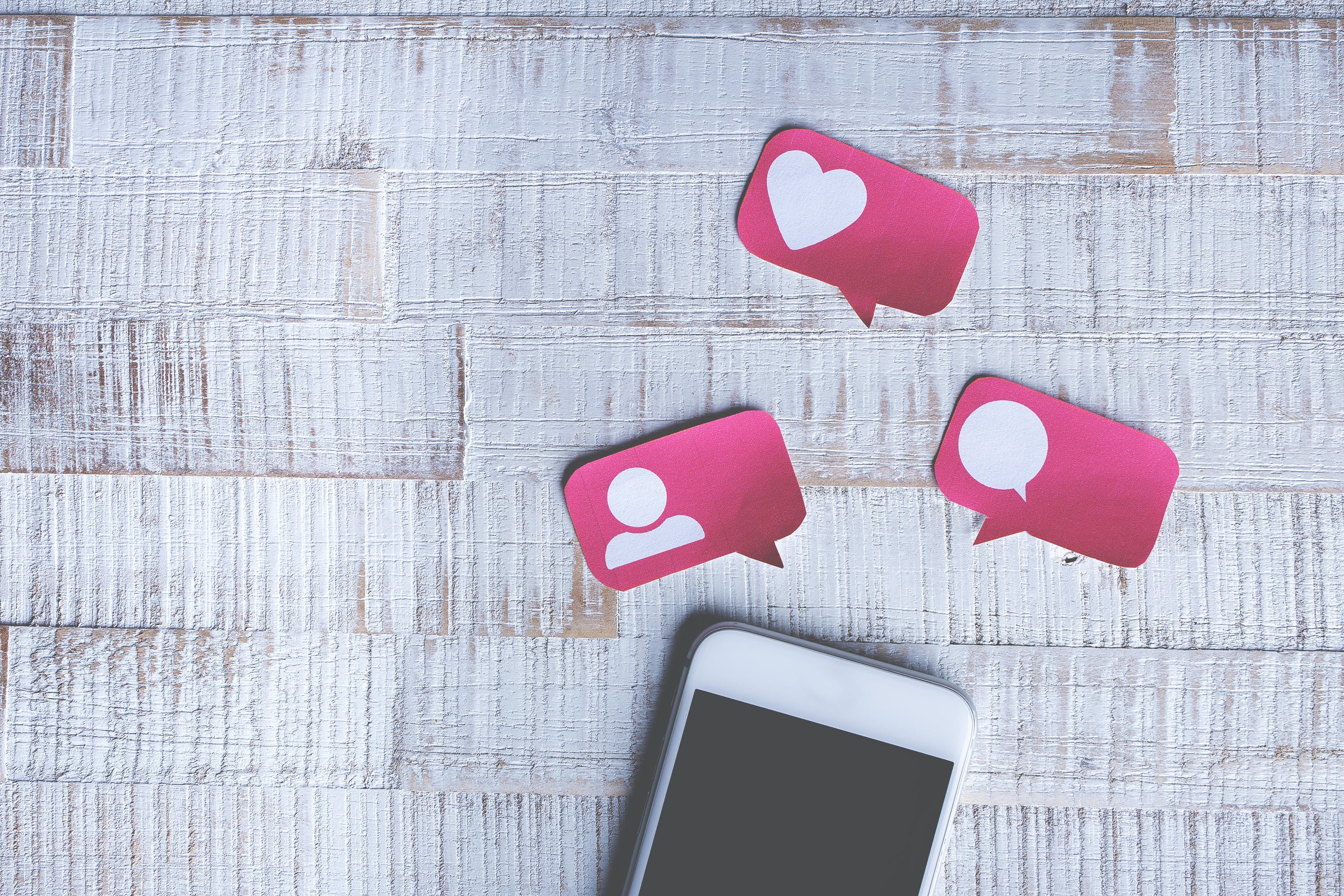 An iPhone on a wooden desk with cardboard symbols for likes, comments and friend requests beside it.