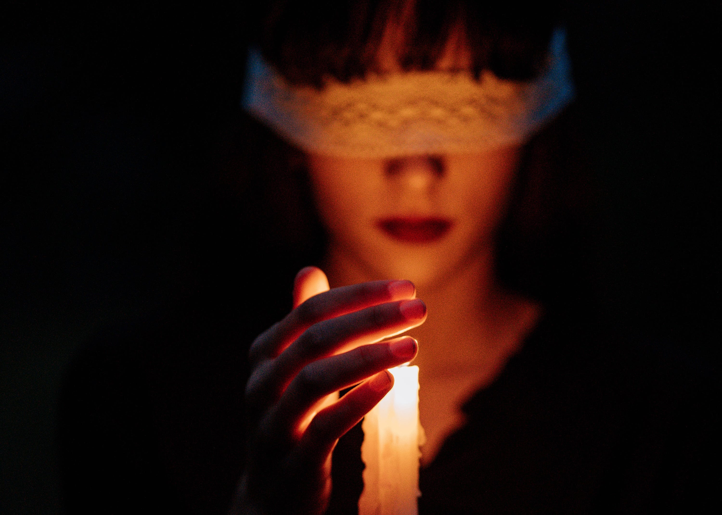 Woman in lace blindfold holding a candle, face softly illuminated in dark room, hypnotic intimate mood
