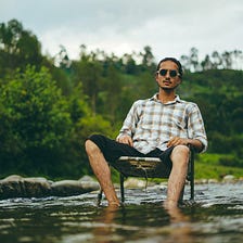 A man practising Loving Kindness Mindfulness sat in a chair in a stream outside in nature.