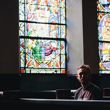 Man sitting in dark church pew beside stained glass window.
