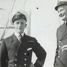 Phot depicts two naval captains standing outside in the sun, in their uniforms, laughing and one is smoking.