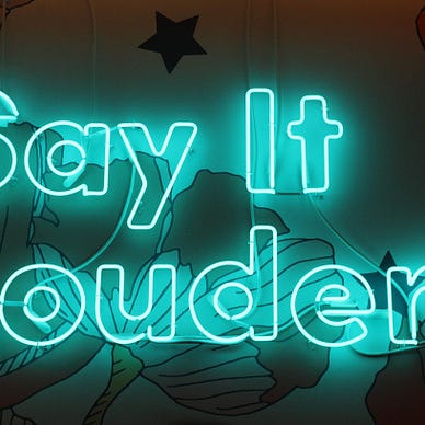 A neon, tube lettering sign which reads “Say it louder”