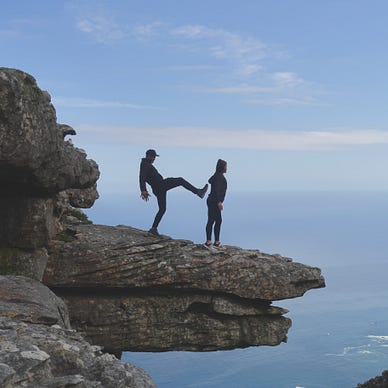 A man pretending to kick a woman in the back while she stands near the edge of a cliff.