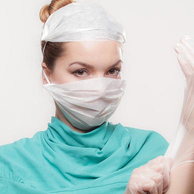 A dark-haired white female doctor glances meaningfully at us. She’s wearing turquoise protective clothing and two white masks. She’s just finished adjusting the fit of a transparent white plastic glove on her left hand.