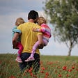 A father walking in a meadow with flowers, holding two young girls, with a young boy walking ahead.