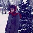 My sister Carol standing in front of a snow dappled Christmas tree, wearing a wool coat and a red scarf.