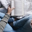 Someone sitting on a bed holding a book open (you can’t see their face). They are sitting cross-legged with ripped jeans and a gray long sleeve shirt.