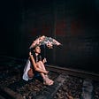 Beautiful young Asian woman sits in the dark on what looks like wide railroad ties, inside a track, looking up pensively into and through a clear umbrella with large, lit stars hanging from it.