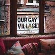 Showing a sofa inside a room where the window looks out on a sign that says, “Our Gay Village — Manchester”.