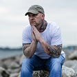 Man sitting outside on a rock looking lost in thought and a bit sad. Pensive. He is wearing a white t-shirt and jeans, with a green ballcap. His arms are tattooed. He has workboots on. The background is blurred but looks like a beach’s rocky shore.
