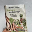 A hand holding up a vintage cookbook with a quaint illustration of fruits and veggies on the front.