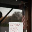 Sign saying closed until further notice on a restaurant door with an American flag reflected in the glass