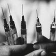 Black-and-white photo: Closeup of a healthcare worker holding five hypodermic needles, syringes unsheathed.