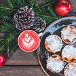 Beautiful sugared tarts rest on a dark-glazed plate on a rustic wood table, next to a red drink in a mug topped with a pretty heart-shaped cream garnish, and a few green bows, pine cones, and red apples.