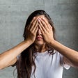 A person with long hair stands in front of a gray background, covering their eyes with their hands.