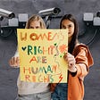 Two women hold a sign saying women’s rights are human rights in front of several sureilance cameras.