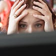 A photo of a stressed woman with her hands on the sides of her face, looking at her computer screen.