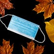 A blue paper surgical mask and brown and yellow leaves on a black background.