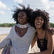 that image is that of two beautiful dark skinned young women taking a picture on the beach,; they are happy and smiling; the world is their oyster; the sand and shoreline are in the background along with the outline of a bluff covered in vegetation.