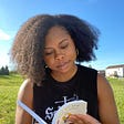 Brianna is staring down at the book she is holding. Their hair is in a wash and go. The background is blue sky and green, freshly cut grass. She is wearing a black muscle top.