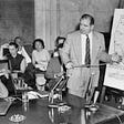 Joseph Welch (sat on the left) and Senator Joseph McCarthy (standing on the right pointing at a diagram) during the 1954 Army-McCarthy hearings. A number of people are sat in the background.