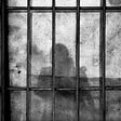 a shadowed figure is seen from behind bars as in jail.