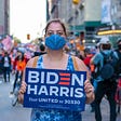 Woman wearing mask holding Biden Harris sign on crowded street.