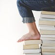 How to Comprehend More in 5 Minutes by Nancy Blackman. Stack of books. Person sitting on books. Feet on top of stack of books. Reading. Writing. Comprehension. Smart Reading Hacks