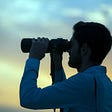 Against a cloudy sunset sky, a silhouette of a man is using a pair of large binoculars. He is wearing a white collared shirt and is implied to be watching something in the distance with the binoculars.