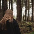 Young girl with black sweatshirt and straight brown hair walking toward heavily forested woods.