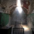 A old prison cell with a bare bed and light entering from above through a tiny window.