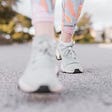A female’s foot just hitting the pavement on a neighborhood walk. She is wearing peach and gray swirly leggings with light gray sneakers.