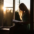 The silhouette of a woman sitting on a windowsill reading a book. Golden sunlight is coming in through the open window.