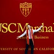 A young male graduate stands in front of a USC Marshall School of Business mural.