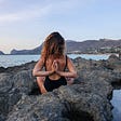 A woman sitting on the rocks looking at the waters and the mountains behind it, her hands are behind her back. 4 things to know ehile starting a spiritual journey.