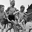 A large group of white tween-aged boys run shirtless. Their shortly cropped haircuts as well as their short swim trunks dub the black and white photo as being from 1950s America. Three boys in the foreground laugh, exposing ribs on their skinny frames. All of the kids are skinny, especially by today’s standards.
