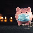 Piggy bank with stacked coins in front and a mask on its face, with out of focus candles in the dark background.