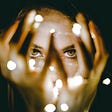 A close up of a woman’s face with her hands in front of it and lights tangled in her fingers.