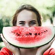 Woman smiling holding a watermelon slice in front of her face.