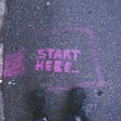 Overhead view of gray concrete road with the words “START HERE…” written in purple chalk.