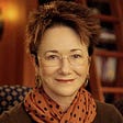 headshot of white woman with short spiky brown hair wearing orange scarf w black polkadots and rimless eyeglasses. big earrings. smiling at camera. she’s sitting in a library with a ladder for reaching books.