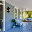 An inviting place to retire on charming blue wrap-around front porch, swing hanging from ceiling, wicker love seat, windows framed in white, black door