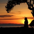Silhouette of a woman sitting on the ground under a tree watching the sun set.