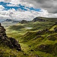 A stunning Scottish landscape shows gently rolling bluffs and valleys covered in emerald grass, beneath a cloudy bright blue sky.
