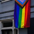 Progressive Pride flag hanging vertically on a blue-gray building