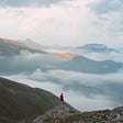 hiker standing on the edge of a cliff overlooking a mountain range above the clouds