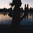 A close up silhouette of a woman with a ponytail runing beside a lake with a city skyline in the background. The sun is setting.