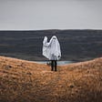 A person standing on a hillside in a sheet like a ghost is waving at the camera.