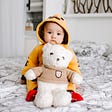 A cute toddler boy sitting on a bed with a white teddy bear in front of him. They boy has a sweet, inquisitive expression.