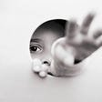 A child’s face partially obscured by a hole in the wall, but their hand reaching out through it towards the camera.